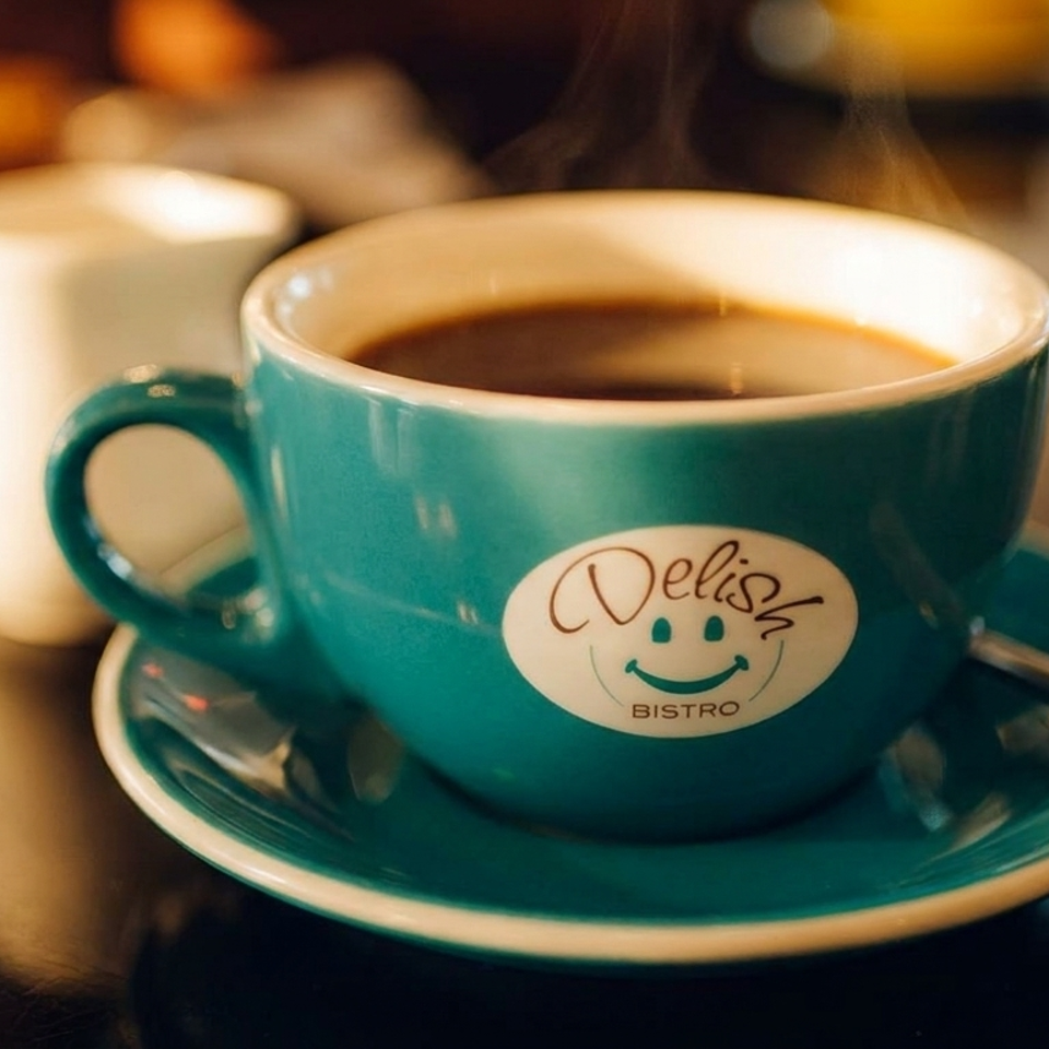 Steaming coffee in a turquoise Delish Bistro cup and saucer with a spoon, beside a white cup on a café table.